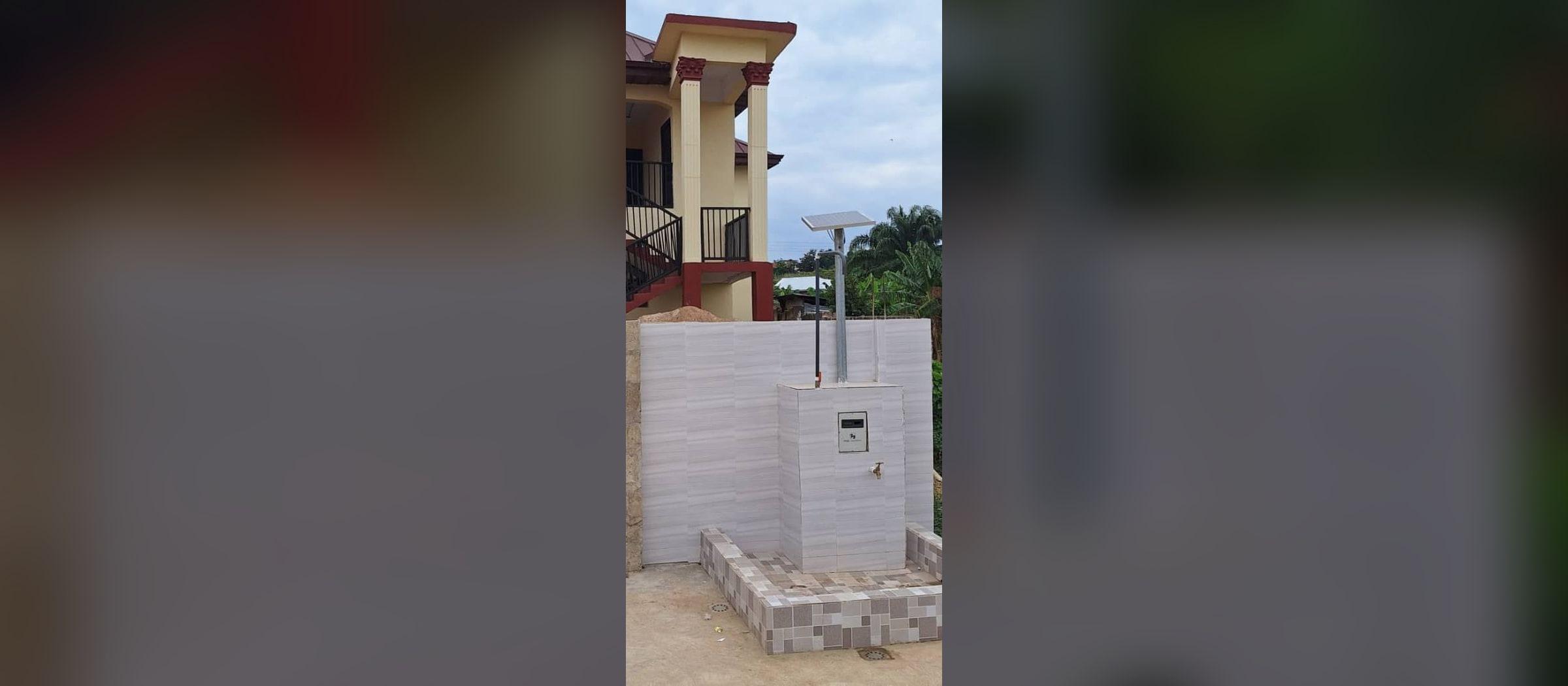 A solar-powered Maji Community Dispenser on a grey-tiled standpipe base in Kumasi, Ghana, in front of a house with red-trimmed columns