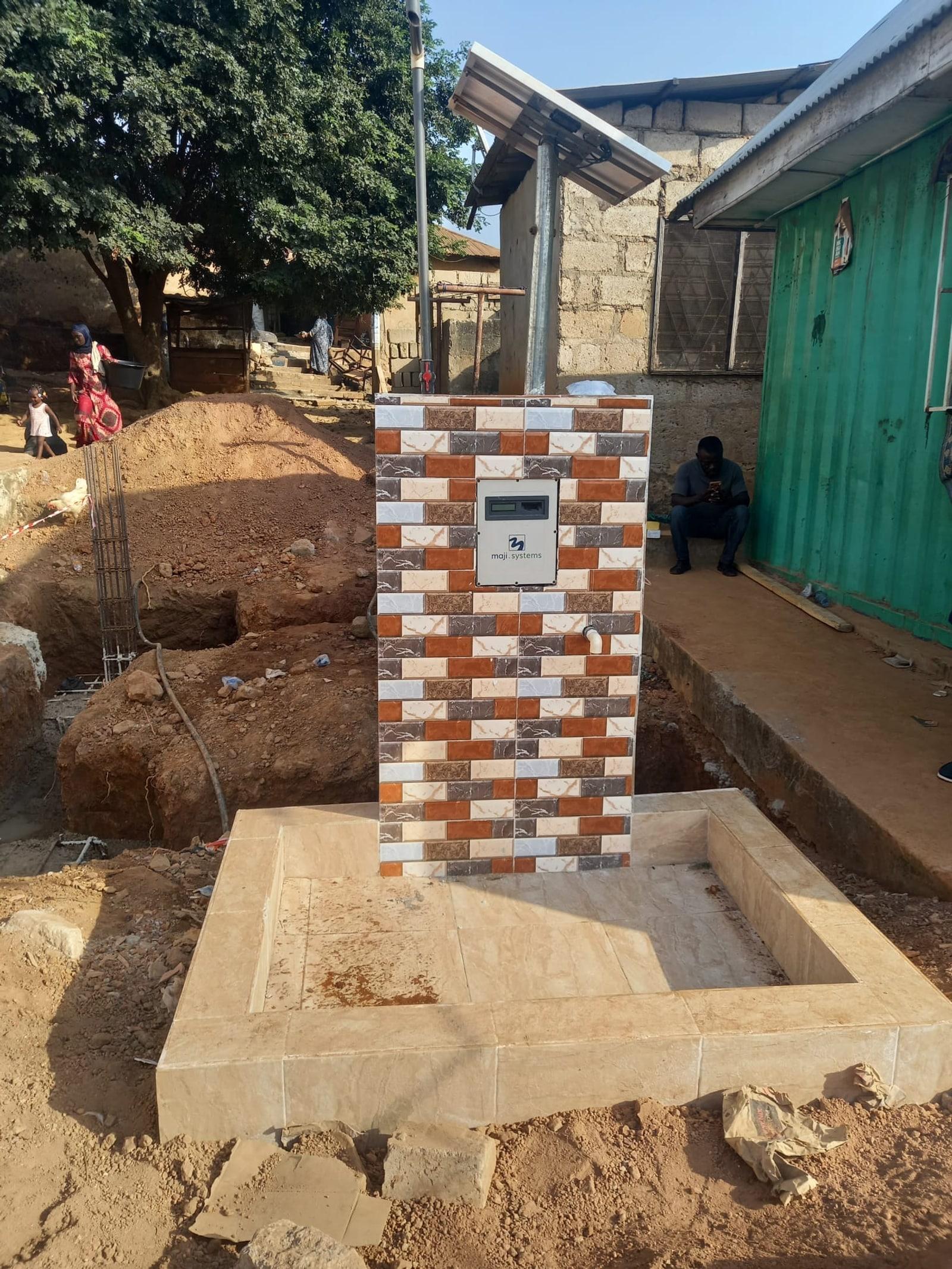 A woman and child passing a solar-powered Maji Community Dispenser standpipe in a Kumasi neighbourhood